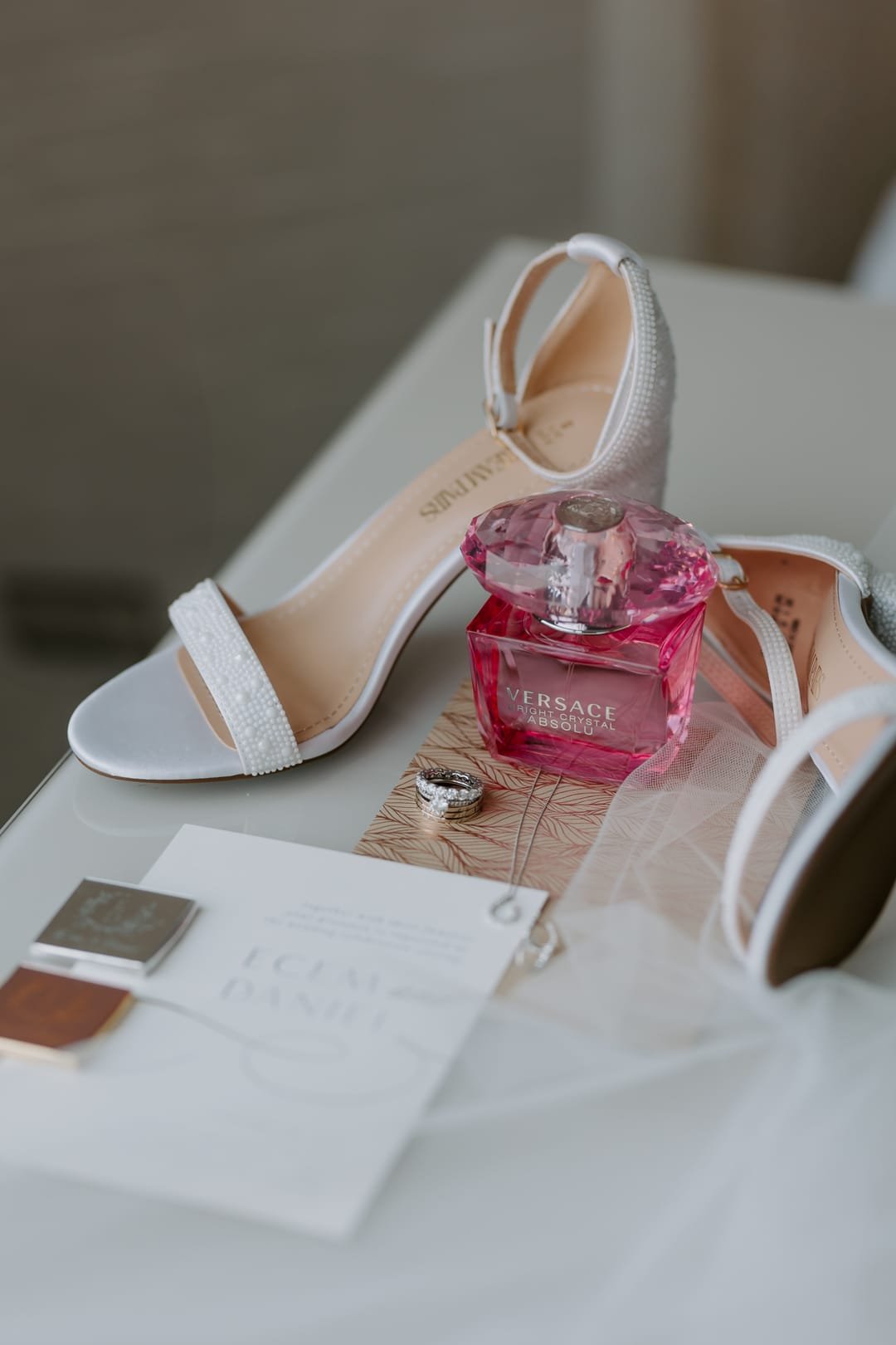 Bridal heels, a pink perfume bottle, the ring and the invitation suite laid out on a white table