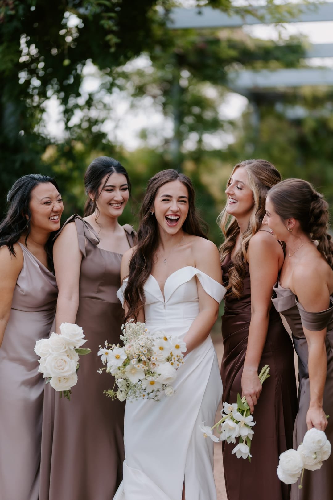 Bride laughing mid-story with five bridesmaids in mocha satin, bouquets at their sides