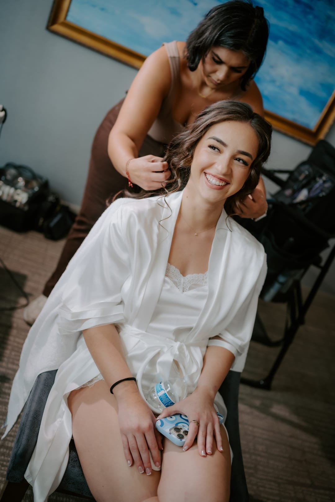 Bride in a white robe smiling over her shoulder while a stylist pins her hair