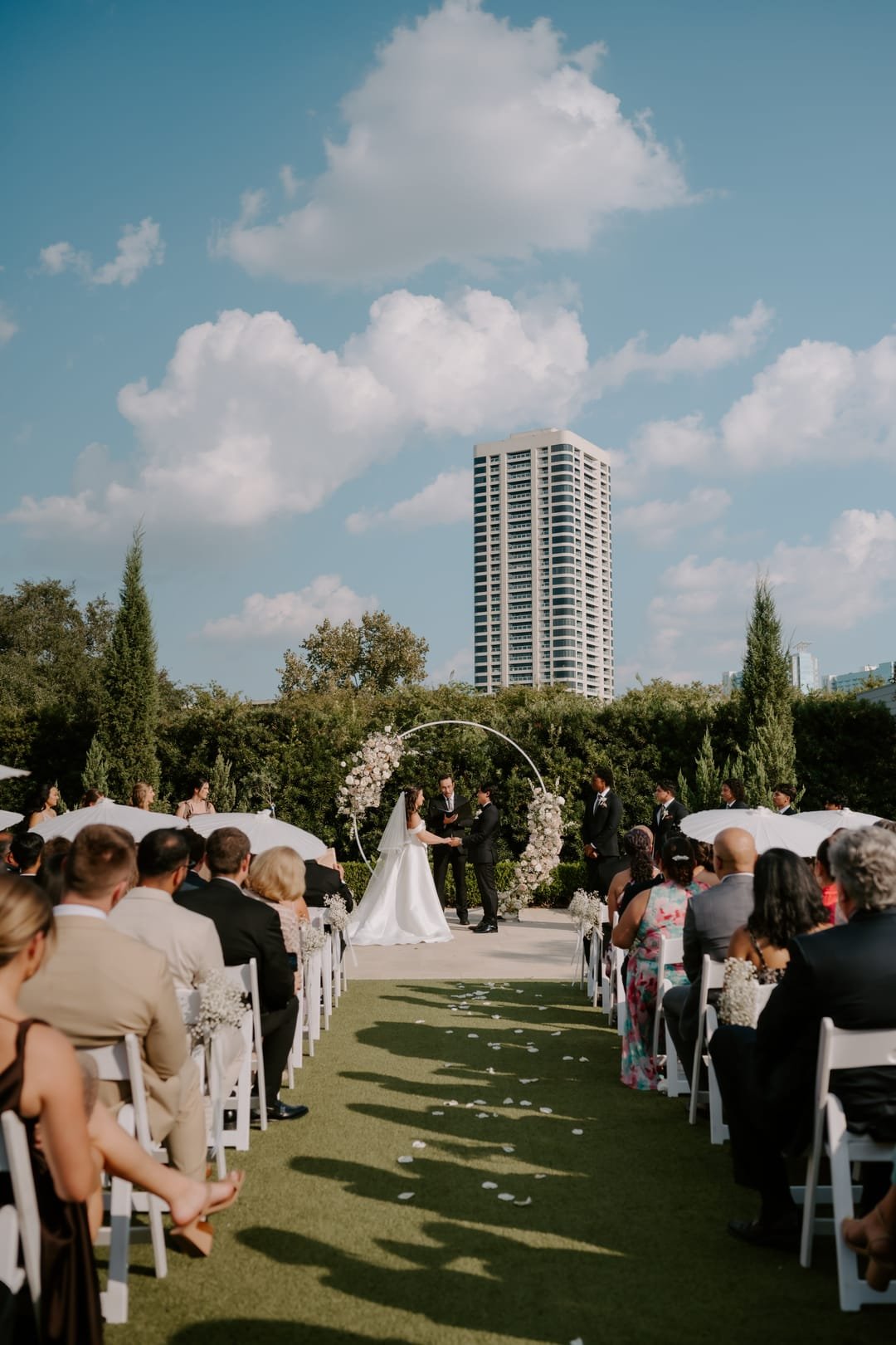 Aisle view of the outdoor ceremony, seated guests either side and a city tower on the skyline