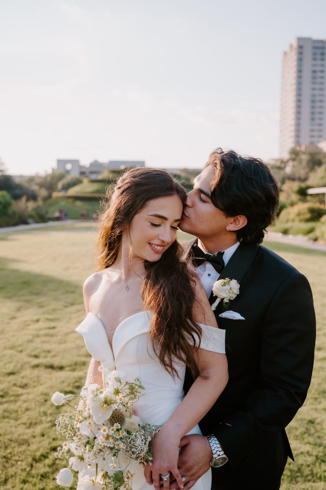 Groom kissing the bride's temple on a sunlit lawn, city towers pale on the skyline behind them