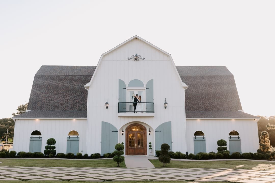 The white barn venue at dusk, the couple standing together on its small balcony