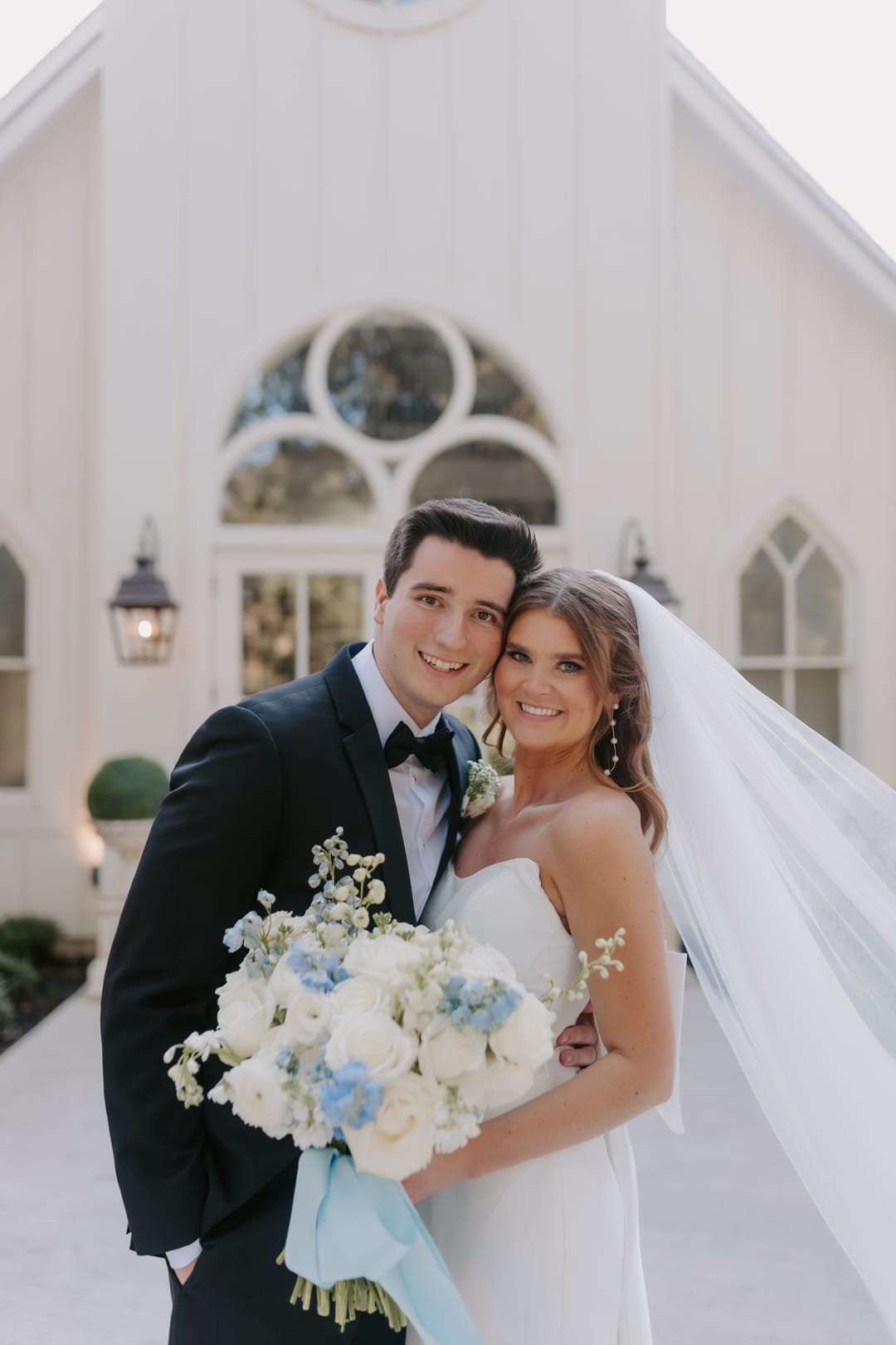 Couple cheek to cheek outside the chapel, her veil catching the light behind them