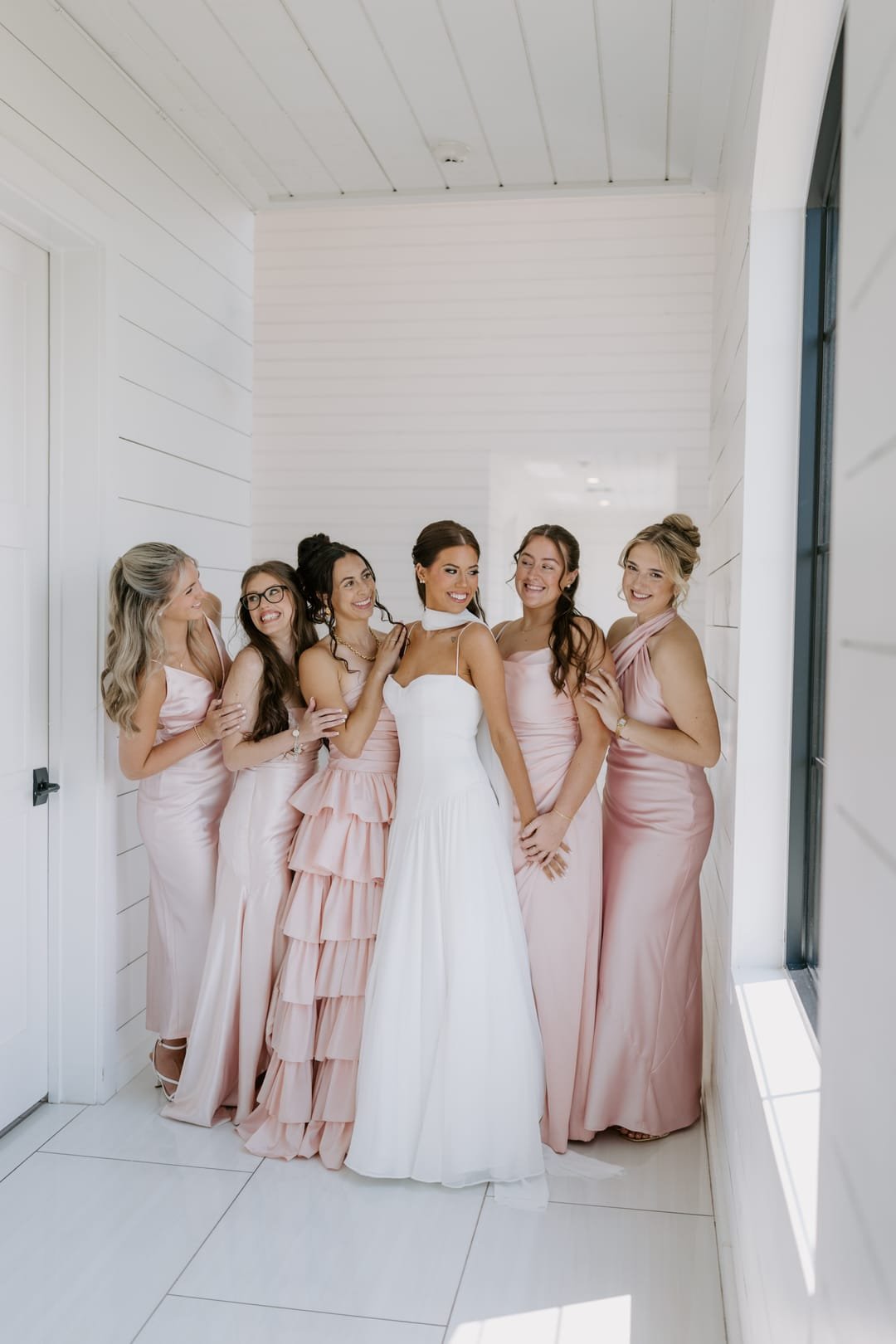 Bride surrounded by bridesmaids in blush gowns in a bright white hallway
