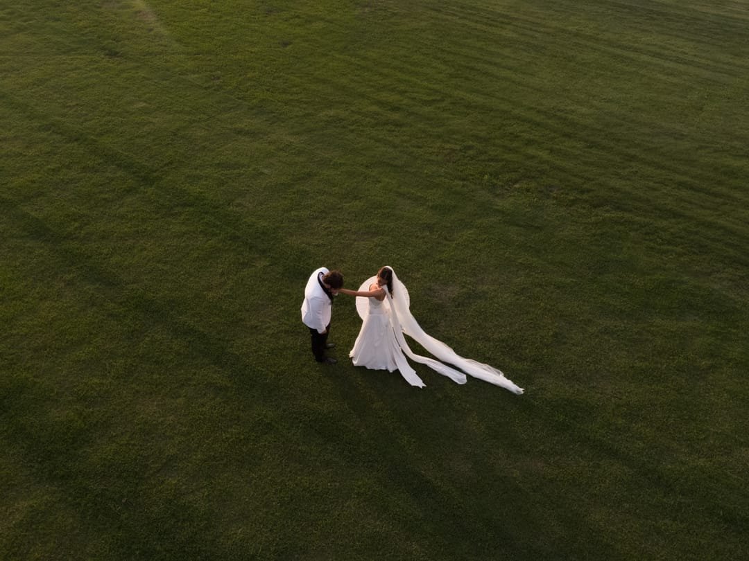 Overhead view of the groom bowing to kiss the bride's hand on a wide green field
