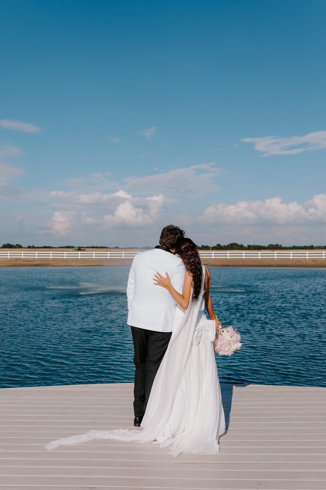 Couple on a dock looking out over the lake, her veil pooled on the boards