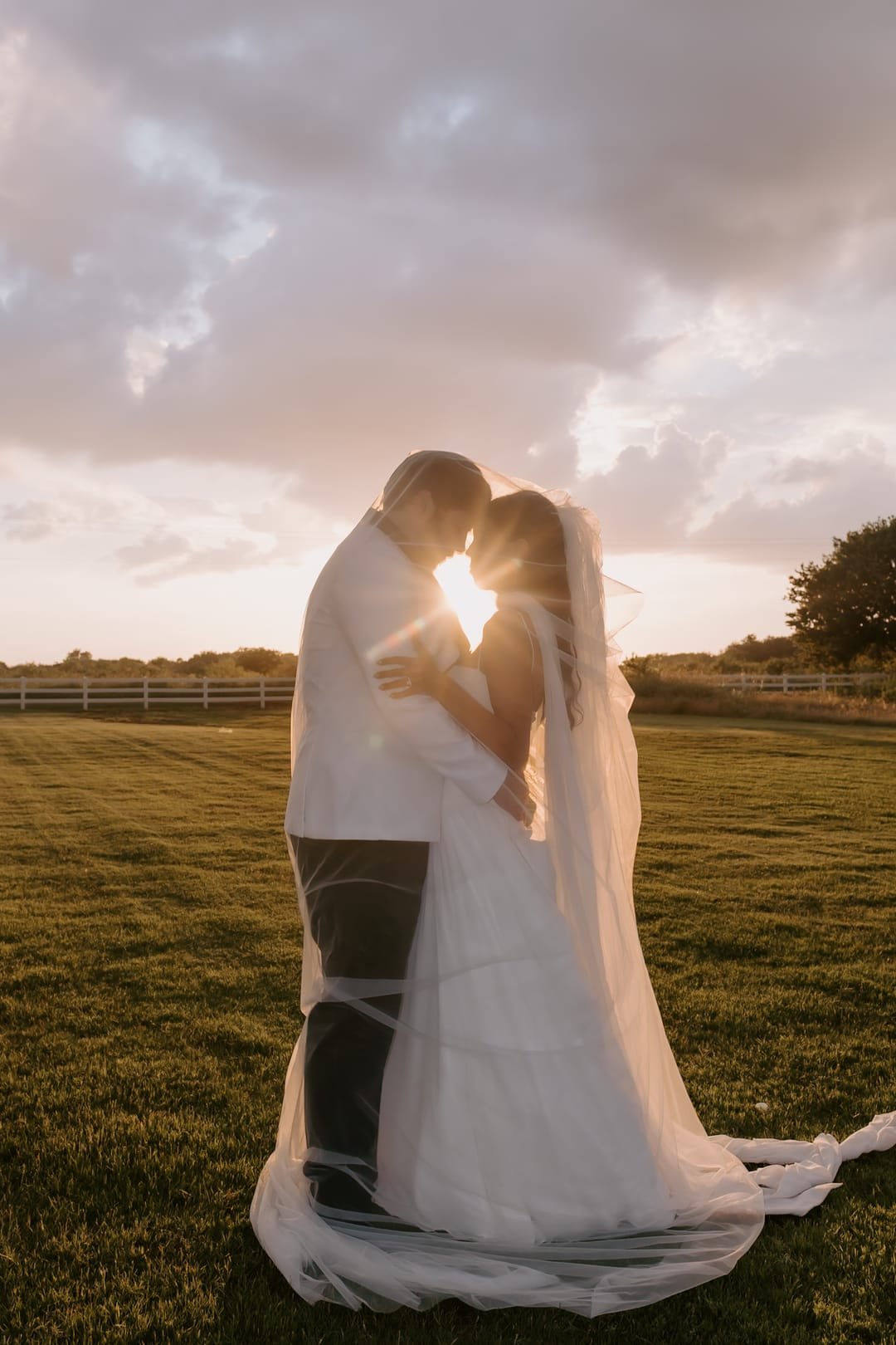 Couple kissing in silhouette as the sun flares between them, veil trailing across the grass